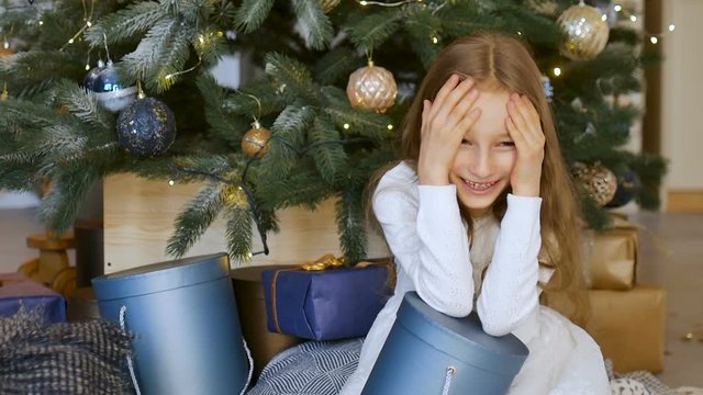 Cute blond girl has recieved a dark blue gift box sitting near christmas tree, happy childhood and xmas celebration concepts