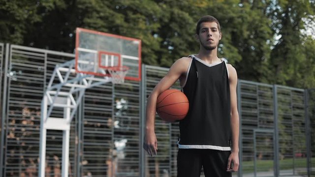 Portrait of Young Guy Basketball Player Wearing Black Singlet Holding Ball Standing at Street Basketbal Court and Looking to Camera. Healthy Lifestyle and Sport Concept.