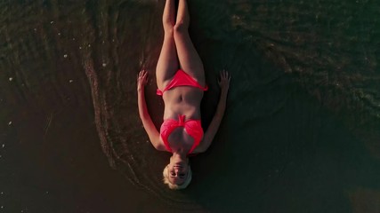AERIAL: Birds View of young confident blonde woman with pink bikini laying flat on beach shore line with waves rushing over her body [4K] - Powered by Adobe