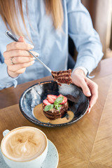 Woman eats chocolate sponge cake, with caramel, decorated with strawberries and mint leaf. Breakfast in the cafe, morning coffee. on wooden table. Restaurant menu