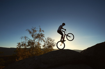 Silhouette of athlete cyclist standing on back wheel on trial bicycle. Sportsman rider balancing on the edge of big boulder on the top of mountain at sunset. Concept of extreme sport active lifestyle