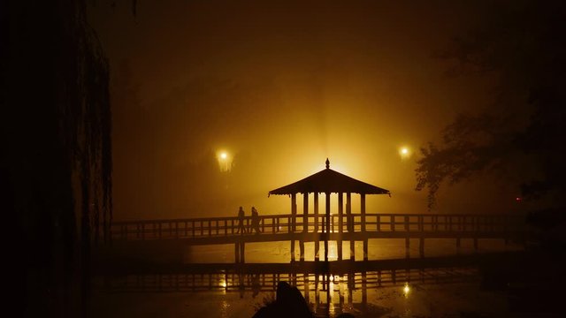 Dreamlike mysterious dark park with a rotunda in japanese style. Silhouette of gropu of people on the bridge at night. Mystic night scene with a house on river with lamp and lights. Walking at night