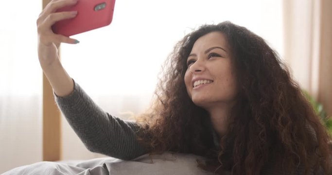 Woman taking selfie using mobile phone camera lying in bed at home