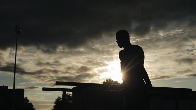 Silhouette of a young man exercising push-ups on parallel bars outdoors. Bottom view. Cloudy sky with little sunshine on the background