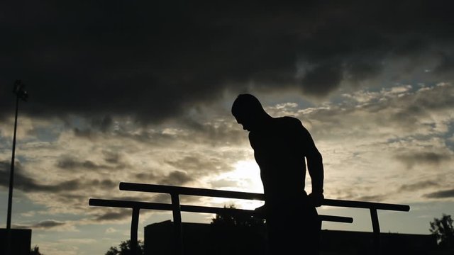 Silhouette of a young man exercising push-ups on parallel bars outdoors. Bottom view. Cloudy sky with little sunshine on the background
