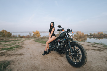 Obraz premium Gorgeous brunette girl with long hair in a black dress and stockings sits and poses on a motorcycle on a background of nature and sky. Portrait of a sexy woman near an expensive black bike.