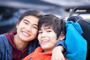 Big sister hugging disabled brother in wheelchair outdoors, smiling