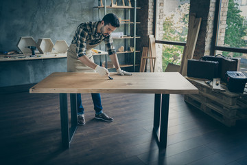 Full length photo of handsome careful guy covering handmade slab table oil varnish paint brush own...