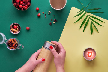 Zero waste tea to go, making herbal infusion in eco friendly insulated bamboo steel flask with herbal mixture and fresh cranberry. Trendy creative flat lay with hands, two tone green and yellow paper.