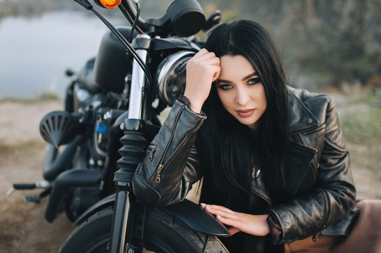 Seductive brunette girl with long hair in a black leather jacket sits near a modern motorcycle on a background of nature. Closeup portrait of a sexy woman near an expensive black bike.