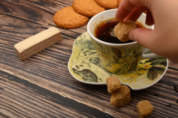 The girl's hand puts a piece of brown sugar in a mug of black tea, oatmeal cookies, tea leaves, waffles, brown sugar on a wooden background. Close up.
