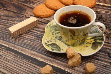 A Cup of black tea, tea leaves, pieces of brown sugar, oatmeal cookies, waffles on a wooden background. Close up.