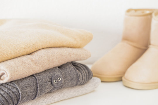 Stack of cosy knitted sweaters in beige grey colour  and beige ugg boots on white lbackground.  Warm comfortable clothes for Autumn Winter season.
