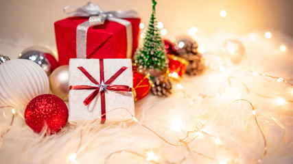 Christmas decoration on white hairy carpet with illuminated lights. Gift box with ribbon, glossy ball, pine cone and Christmas tree on fluffy rug. Present and decorative objects for New Year holiday