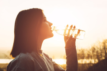 Beautiful female in sportswear drinks water from a bottle at sunset