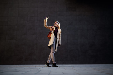 Full length of attractive caucasian smiling blonde woman walking away and waving to a friend. In background is dark wall.
