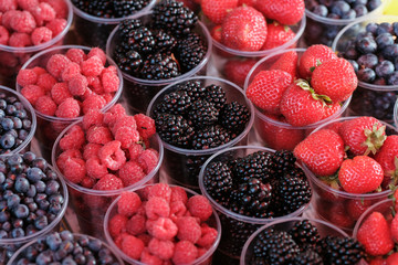 A counter with plastic containers and glasses filled with fresh berries. Blackberry, Strawberry, Blueberry, Raspberry. Food market.