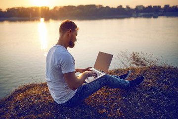 Rear view of handsome bearded caucasian blond man sitting on cliff and typing on laptop. In background is river or lake and sunset.