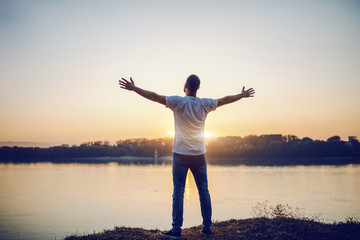 Rear view of handsome caucasian man standing with opened arms on cliff and looking at river and beautiful sunset.