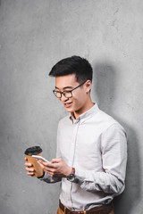 smiling asian businessman holding paper cup and using smartphone
