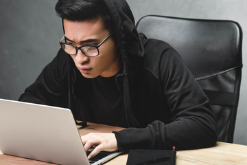 asian hacker in glasses using laptop and sitting at table