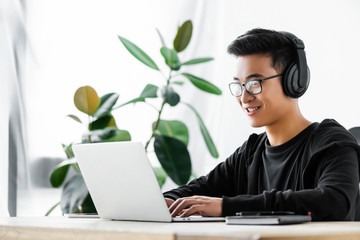 smiling asian hacker in headphones using laptop and sitting at table