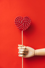 Heart shaped wooden hand holding a large red and white striped lollipop. The concept of Valentine's Day, love and friendship. Minimal simple flat lay composition layout. Natural material and light