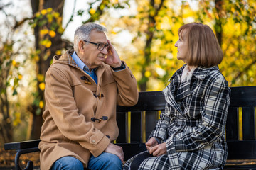 Senior couple is sitting on bench in park and talking about their health problems.