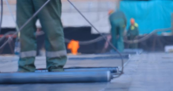 The workers heats the roll of ruberoid with the flame of a gas burner and firmly connect with a surface of stadium main field that needs to be isolated from water