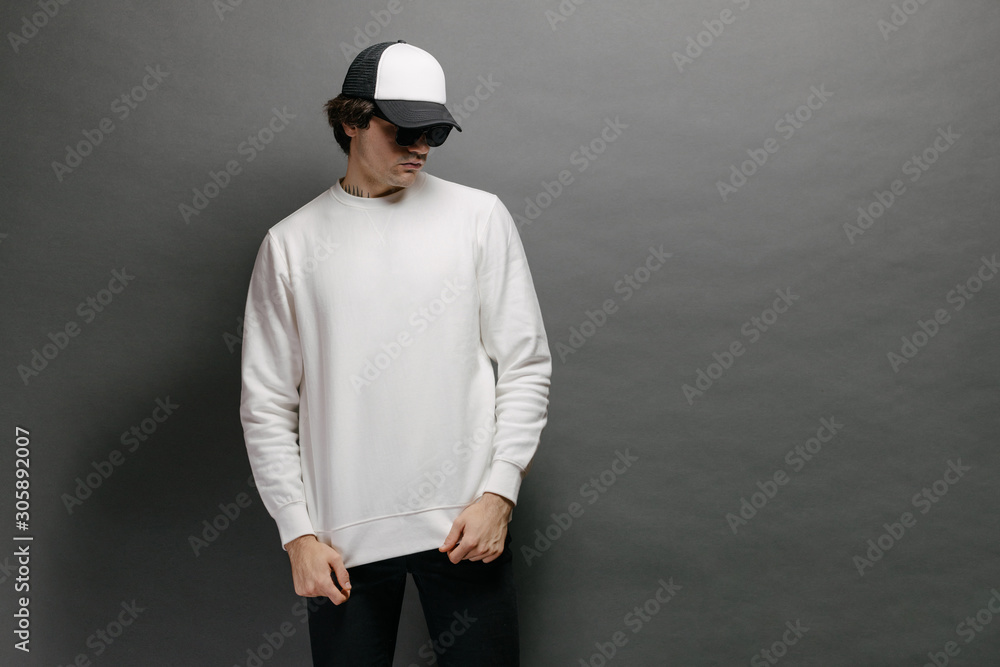 Wall mural Man wearing blank white sweatshirt and empty baseball cap standing over gray background. Sweatshirt or hoodie for mock up, logo designs or design print with free space. - Wall murals