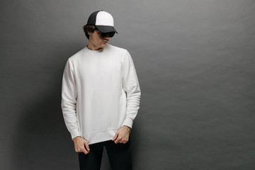 Man wearing blank white sweatshirt and empty baseball cap standing over gray background. Sweatshirt or hoodie for mock up, logo designs or design print with free space.