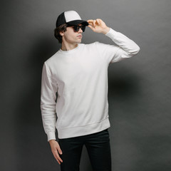 Man wearing blank white sweatshirt and empty baseball cap standing over gray background. Sweatshirt or hoodie for mock up, logo designs or design print with free space.