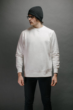 Man Wearing White Sweatshirt And A Black Hat Standing Over Gray Background. Sweatshirt Or Hoodie For Mock Up, Logo Designs Or Design Prints With Free Space.