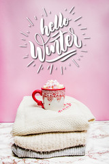 Red holiday mug with marshmallows, sugar cane on a marble surface with pink background with quote Hello Winter 
