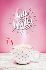 Pink white mug with marshmallows, sugar cane on a marble surface with pink background with the quote Hello Winter