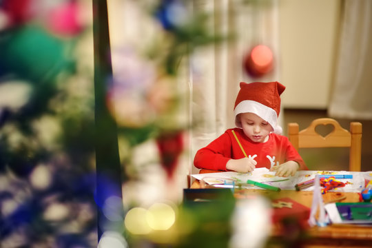 Little boy wearing in a Santa costume is drawing near a decorated Christmas tree.