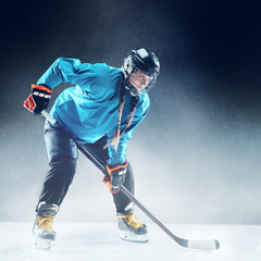 Young female hockey player with the stick on ice court and blue background. Sportswoman wearing...