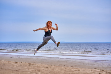 Fototapeta na wymiar A middle aged woman with red hair jumps while jogging. A woman practices gymnastics on the sandy bank of a large river. Cloudy spring morning.
