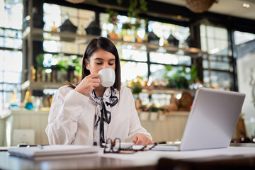 Young caucasian businesswoman sitting in cafe, taking a break from work and drinking fresh coffee. On table are laptop and paperwork.