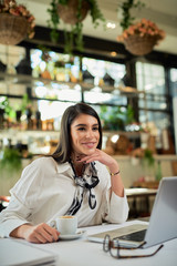 Attractive caucasian elegant brunette sitting in cafe, holdingcoffee and looking at away. On table is laptop.