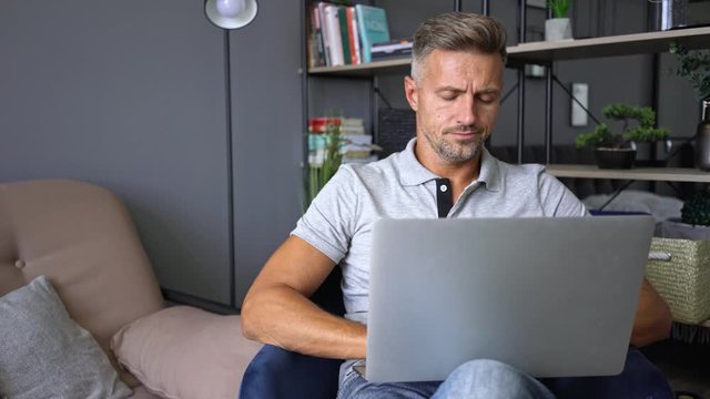 Attractive focused man in casual clothing using his laptop while sitting on armchair in an apartment