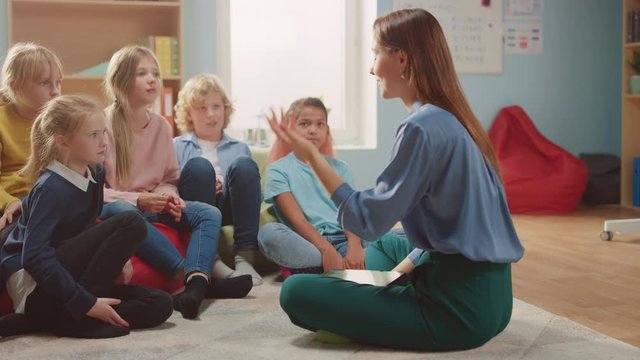 Elementary School Creativity Class: Children Sitting on Carpet while Caring Teacher Explains Lesson while Using Digital Tablet Computer. Children Learning in Friendly Modern Environment. Slow Motion