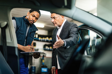 African American mechanic and his customer examining car in auto repair shop.