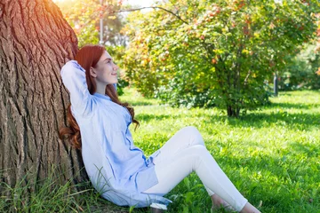 Gordijnen Young woman sitting under tree in summer park © adam121