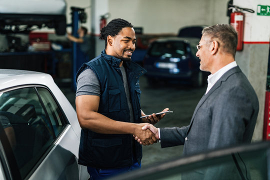 Happy black auto mechanic shaking hands with satisfied customer in repair shop.