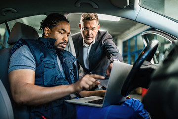 African American auto mechanic and his customer cooperating while using laptop in repair shop.