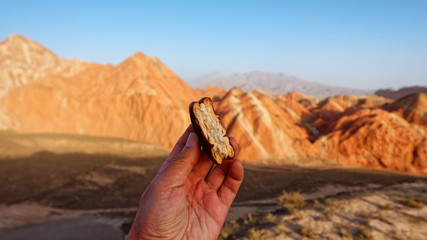 Chocolate biscuit stuffed with bite in hand comparing with Amazing scenery of Rainbow mountain in Zhangye Danxia National Geopark, Gansu, China.