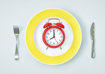 red alarm clock on white-yellow plate with fork and knife on white background. Meal time concept.