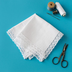 Needlework. Two white handkerchiefs with lace trim, spools of thread, scissors, needles and a thimble on a blue background. Selective focus.