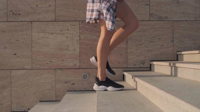 The feet of a young woman in black sneakers, shorts and a plaid shirt climb the stairs, a stone wall in the background. Slow motion. Tracking camera on the object.